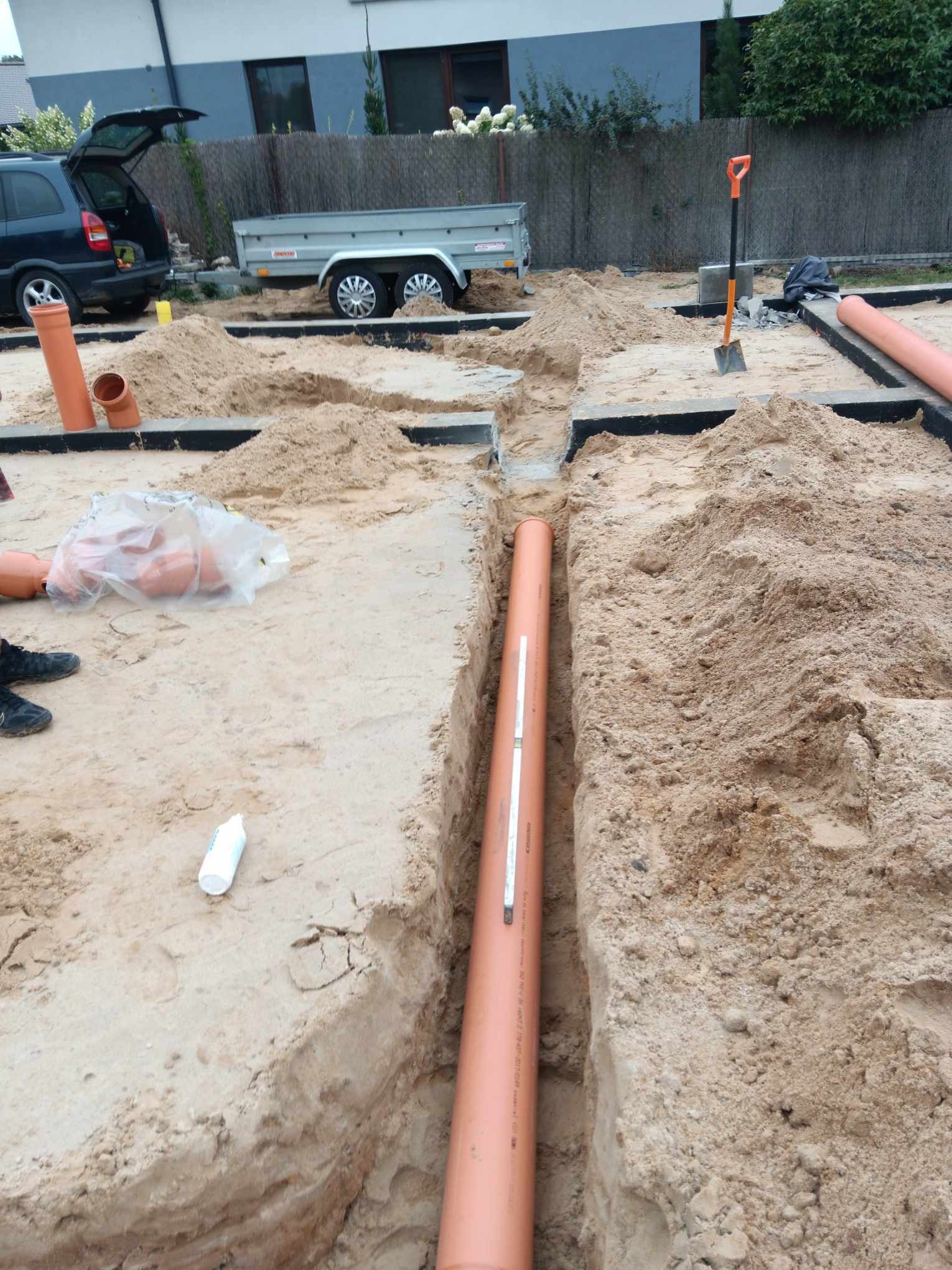 Wykonane wykopy pod instalację rur kanalizacyjnych z widocznymi fragmentami fundamentów, samochodem z przyczepką i narzędziami w tle