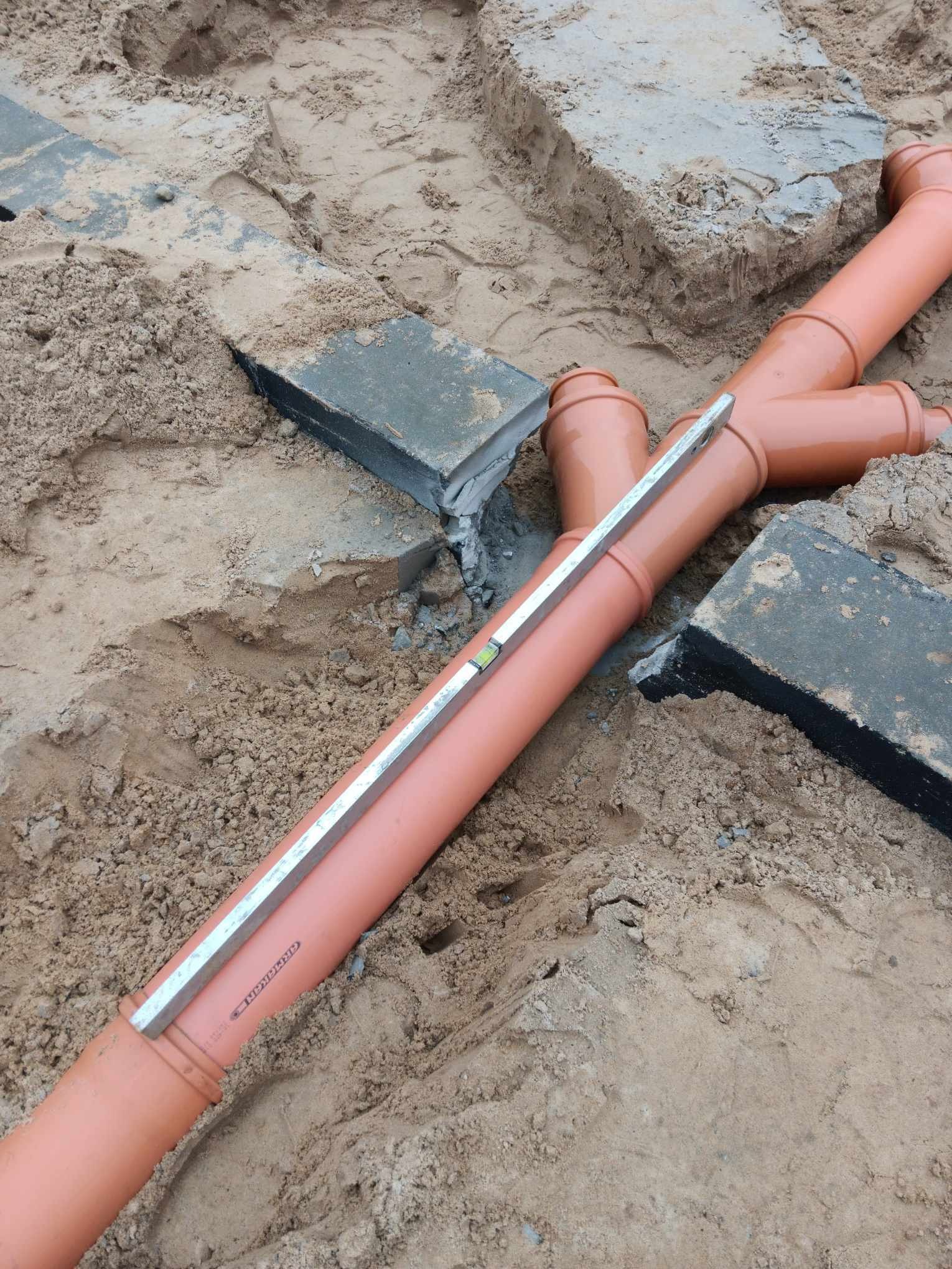 Pomarańczowe rury kanalizacyjne ułożone w wykopie z piasku, poziomowane poziomicą, widoczne betonowe bloki.