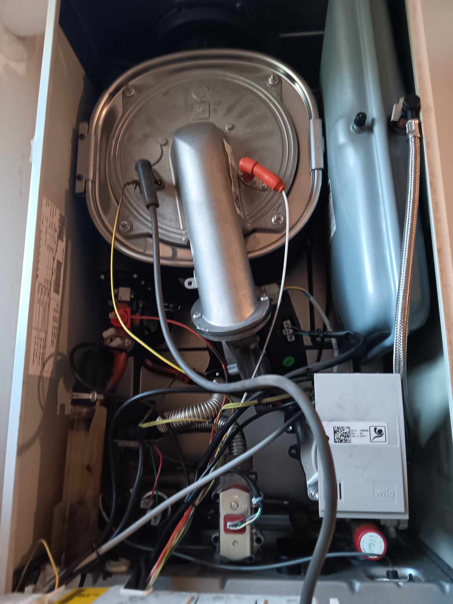 Wnętrze kotła gazowego z widocznymi elementami instalacji, w tym srebrną obudową palnika i zbiornikiem wyrównawczym, liczne przewody i kable wewnątrz urządzenia.