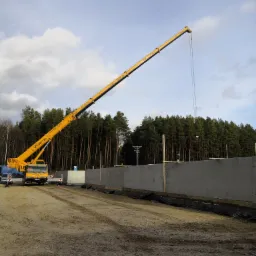 Żółty dźwig teleskopowy z wysuniętym ramieniem na tle betonowej ściany w trakcie budowy, widoczny las i pochmurne niebo.