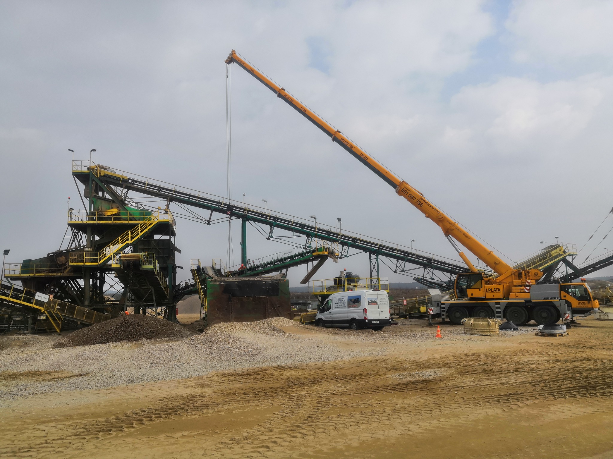Żółty dźwig Liebherr z wysięgnikiem nad instalacją do kruszenia kamienia i transportu taśmowego, obok biały bus firmy I.PLATA z numerem telefonu.