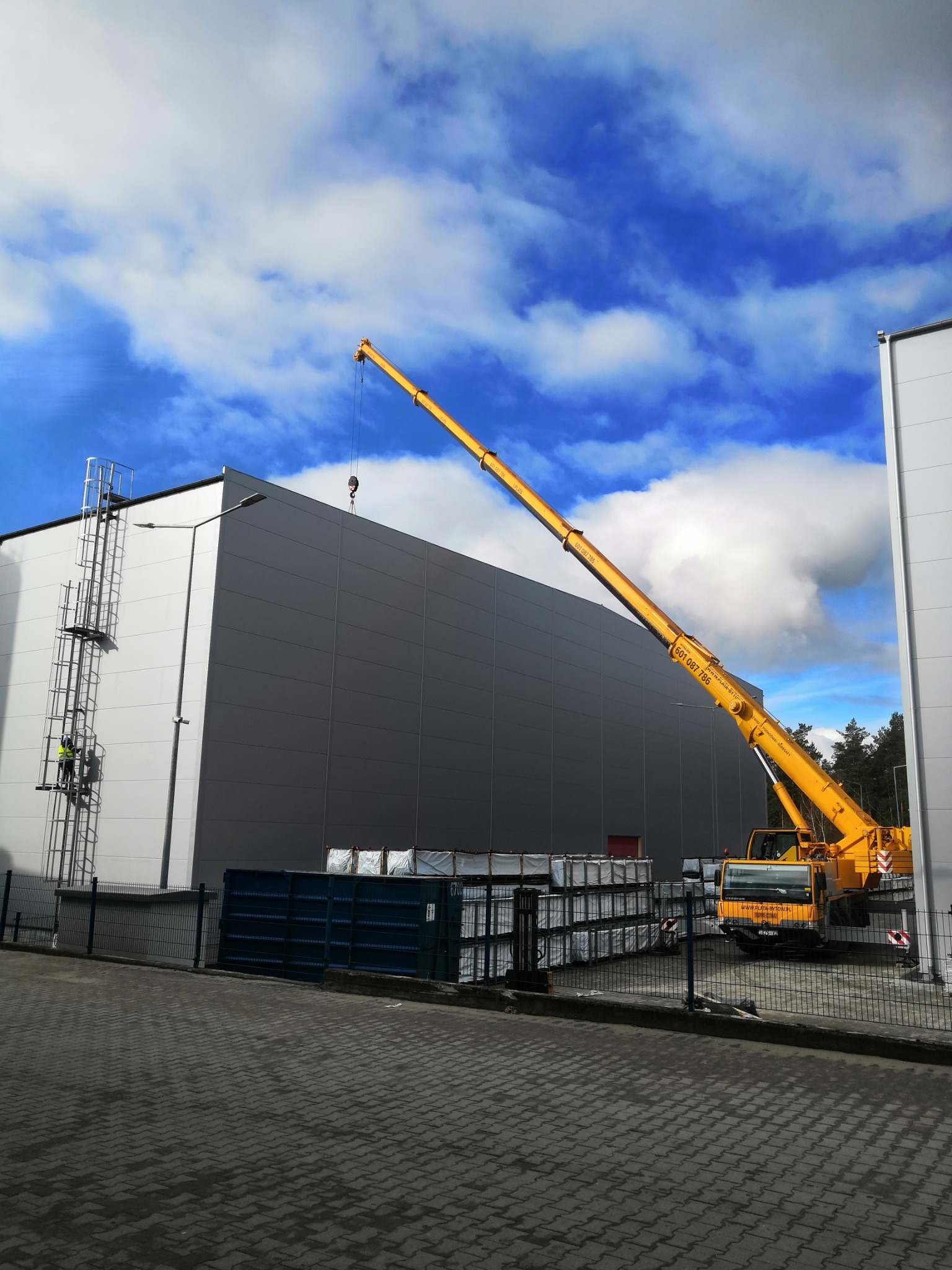 Żółty dźwig samochodowy w Bytowie, z wysięgnikiem skierowanym w stronę szczytu hali, pracownik na drabinie przymocowanej do ściany budynku.