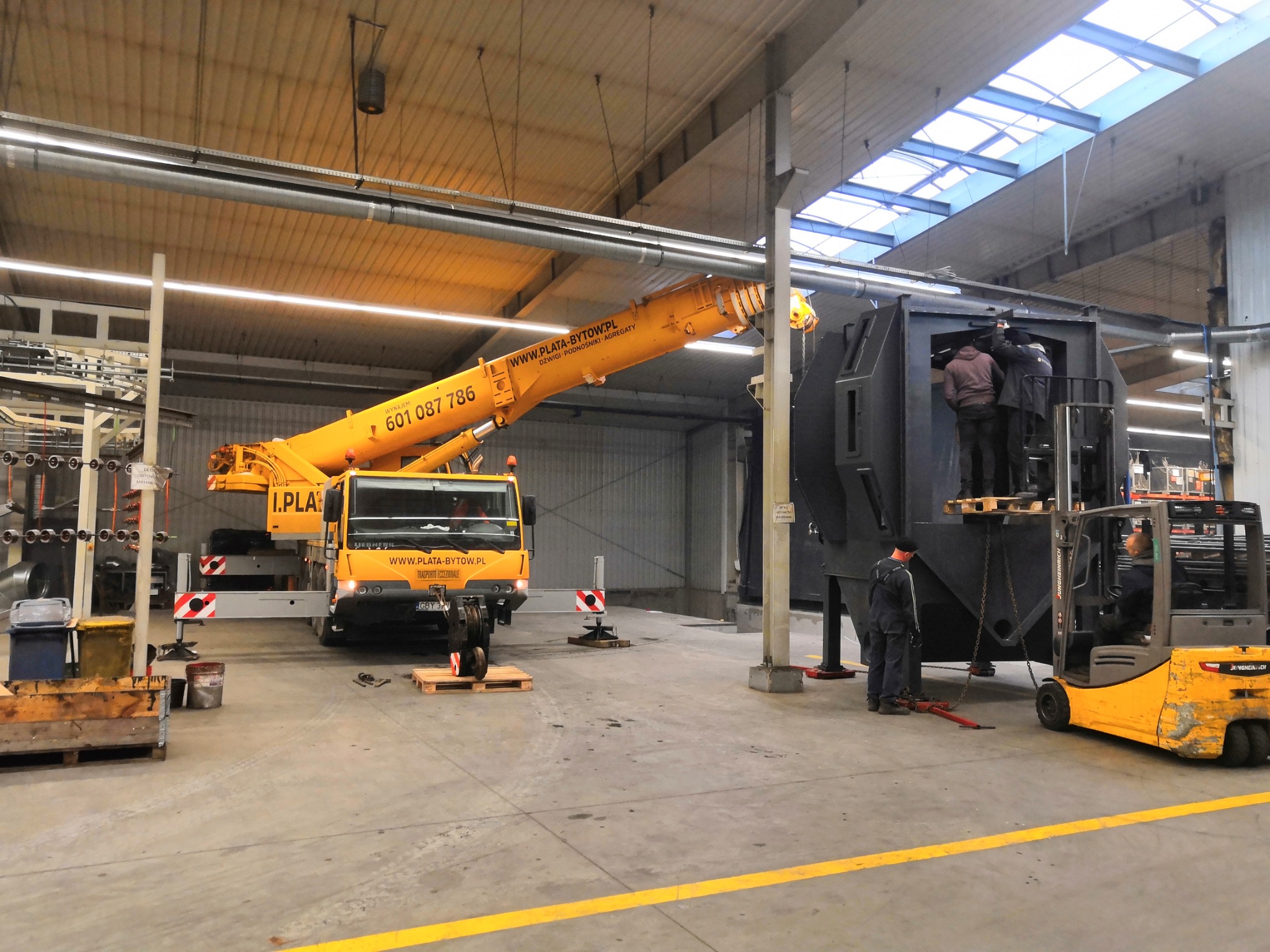 Żółty dźwig samochodowy marki Liebherr, z wysuniętym ramieniem, wspiera montaż dużej, czarnej konstrukcji w hali przemysłowej, gdzie pracownicy na podnośniku widłowym Jungheinrich nadzorują proces.