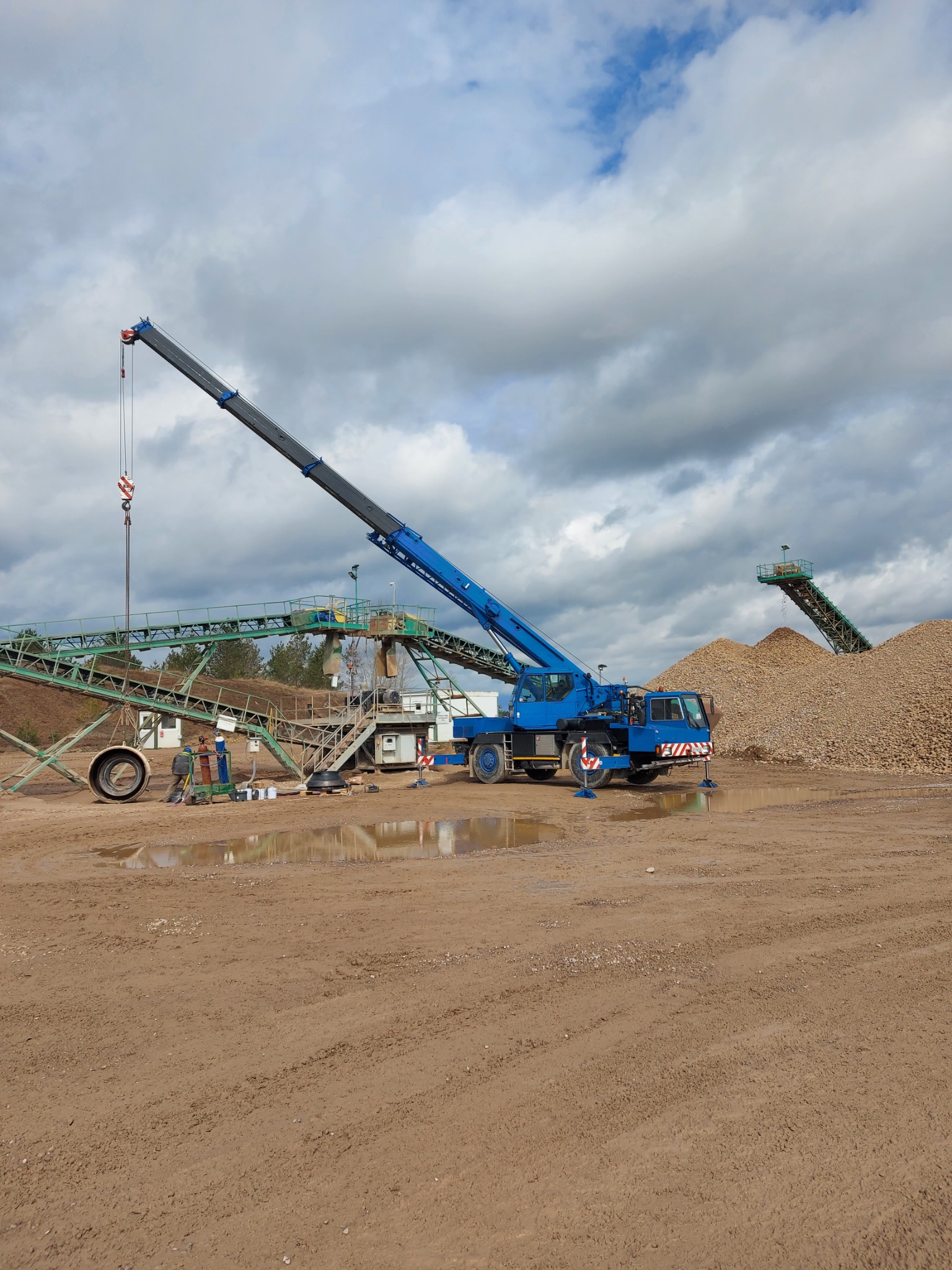 Niebieski dźwig samochodowy w trakcie naprawy przenośnika taśmowego na terenie żwirowni; dwóch pracowników w kombinezonach roboczych przy zielonej konstrukcji przenośnika.