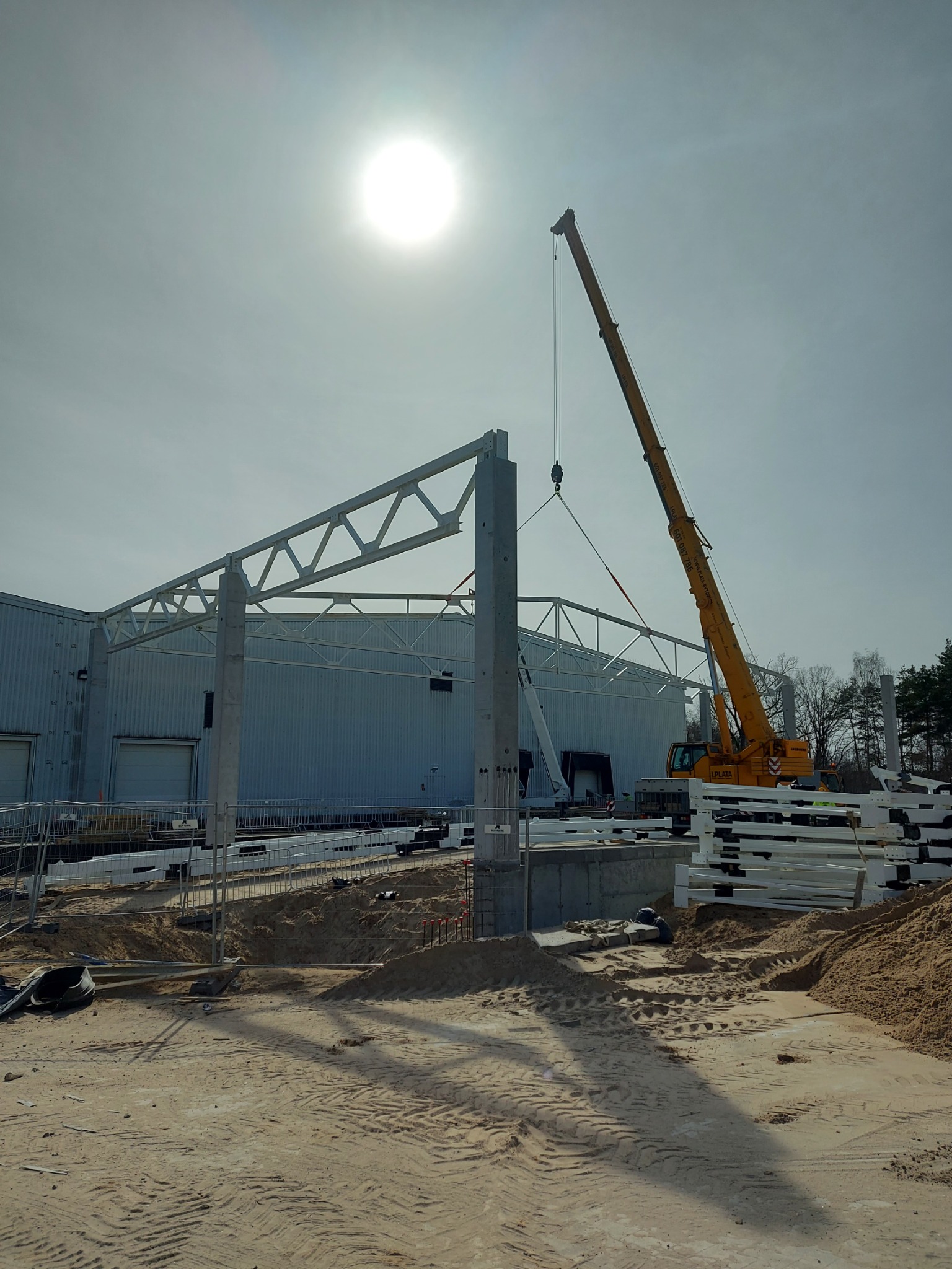 Żółty dźwig podnosi element konstrukcyjny hali magazynowej w słoneczny dzień, widoczne stalowe elementy konstrukcji i teren budowy.