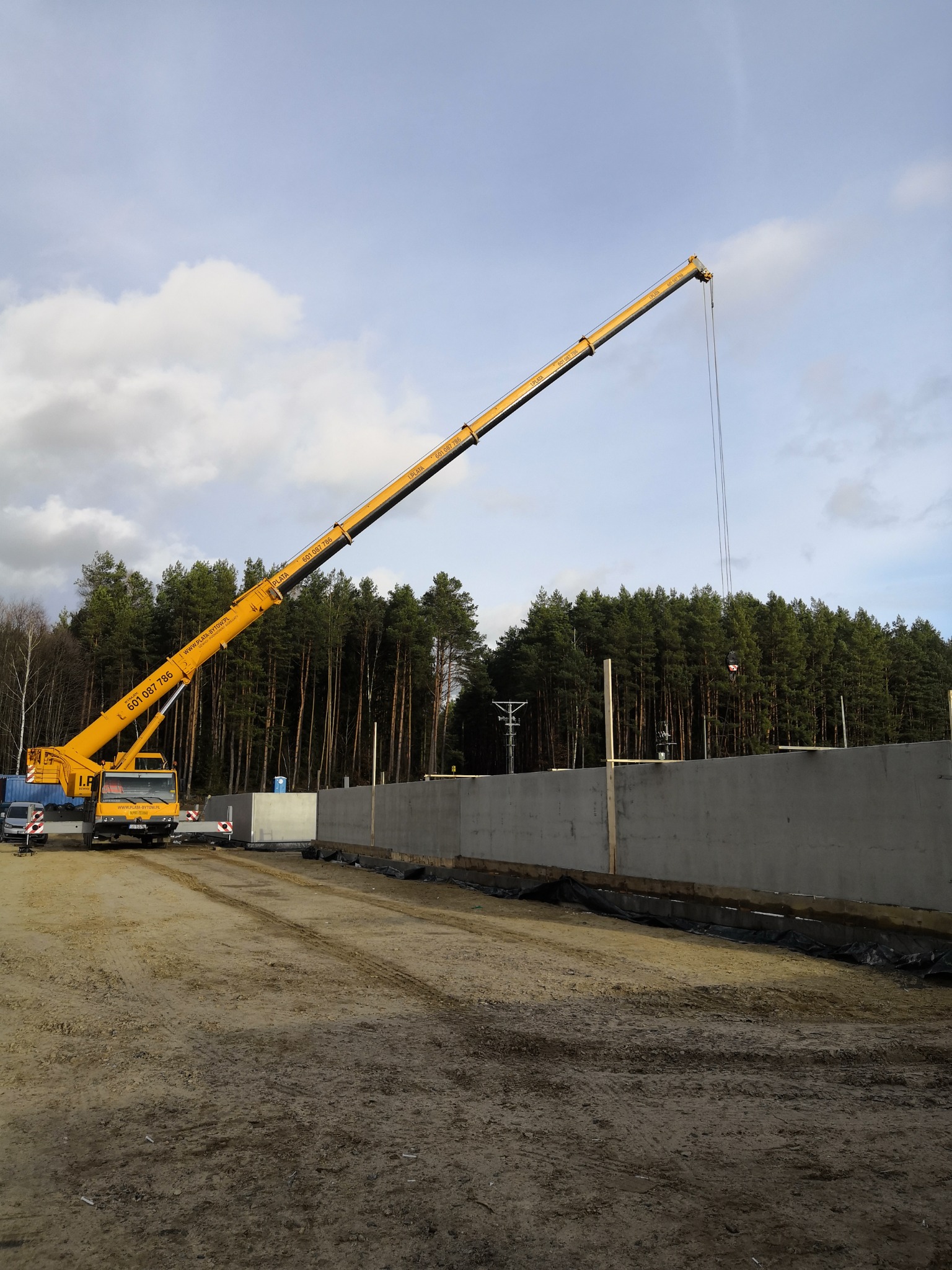 Żółty dźwig teleskopowy z wysuniętym ramieniem na tle betonowej ściany w trakcie budowy, widoczny las i pochmurne niebo.