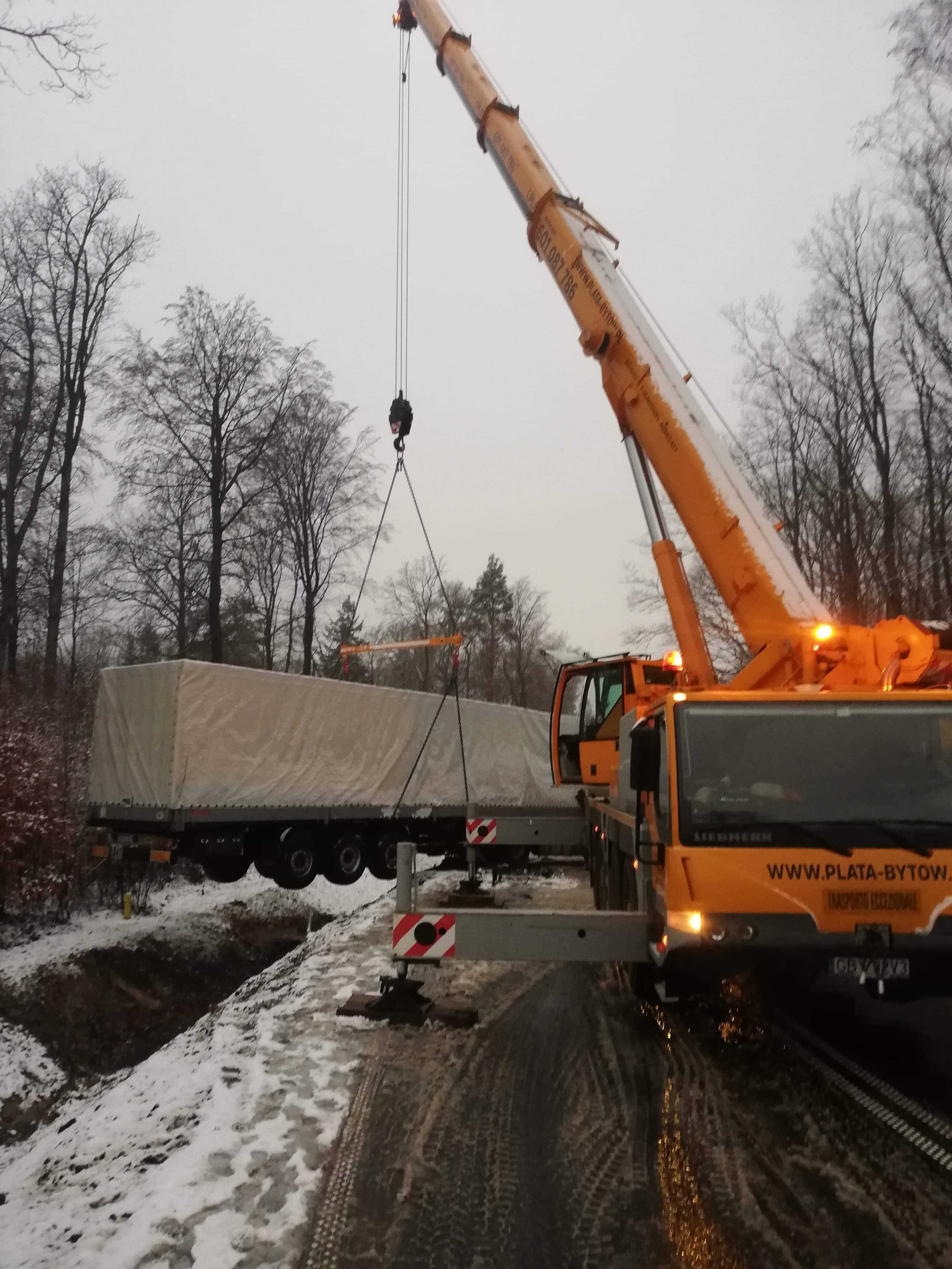 Żuraw Liebherr w zimowej scenerii podnosi przykrytą plandeką naczepę ciężarową, widoczne podpory stabilizujące żuraw i ośnieżone drzewa w tle.
