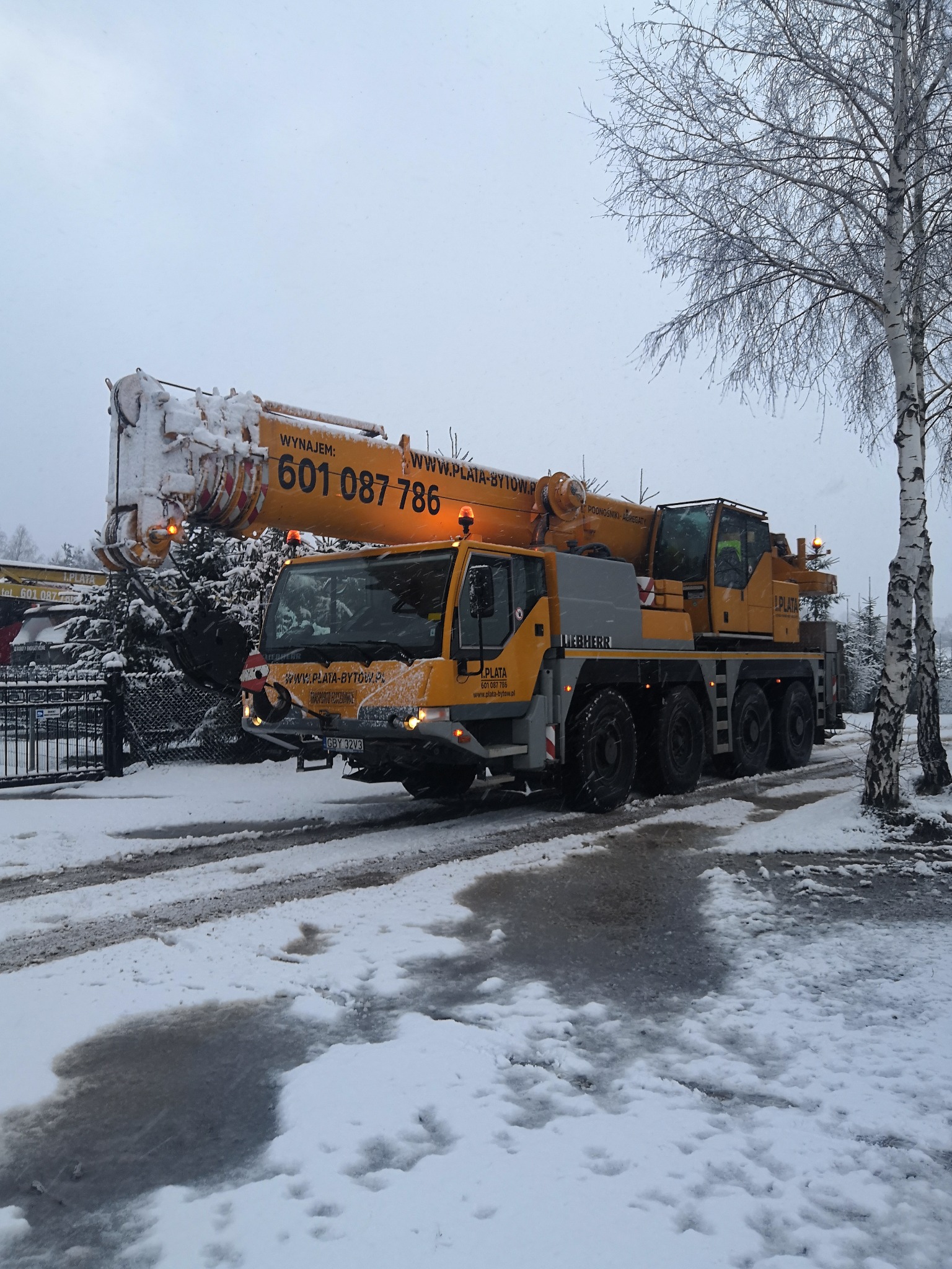 Żółto-szary dźwig marki Liebherr, należący do firmy Plata, z numerem telefonu 601 087 786, stoi na ośnieżonej drodze w zimowy dzień. Na ramieniu dźwigu i na pojeździe leży śnieg. W tle widoczne...