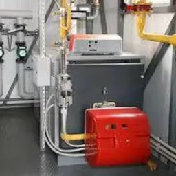 Węzeł cieplny z kotłem, rurami, zaworami i manometrem w pomieszczeniu technicznym, widoczne liczne przewody i elementy instalacji w kolorach szarym, żółtym i czerwonym.