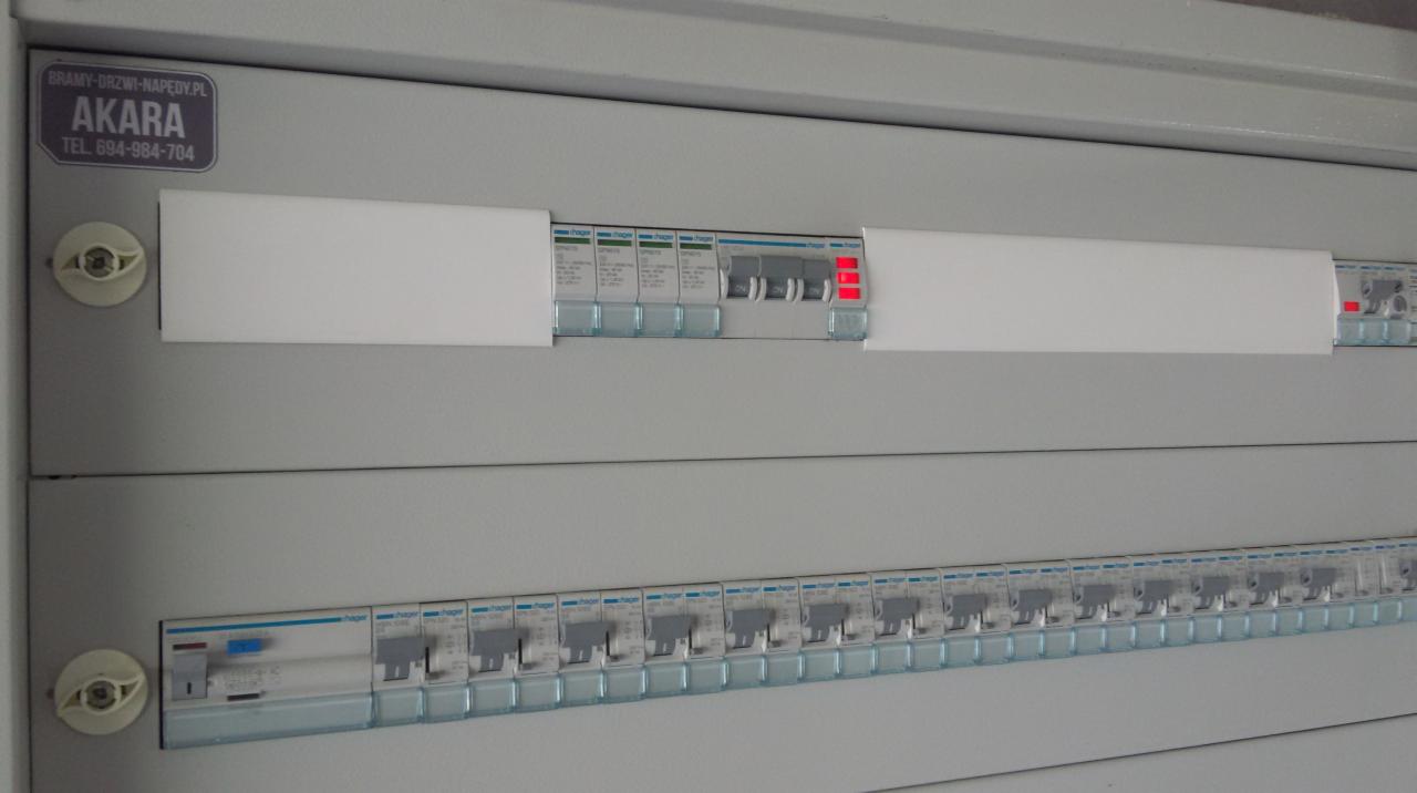 Szara skrzynka rozdzielcza z bezpiecznikami Hager, trzy z nich z czerwonymi diodami, naklejka z logo firmy Bramy-Drzwi-Napędy.pl i numerem telefonu.