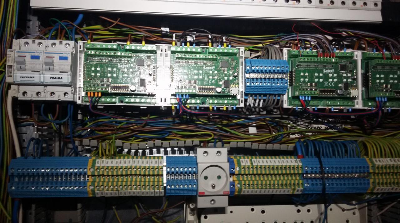 Rozdzielnica elektryczna z modułami sterującymi, bezpiecznikami i licznymi przewodami w różnych kolorach, widoczne złącza śrubowe i gniazdo elektryczne.