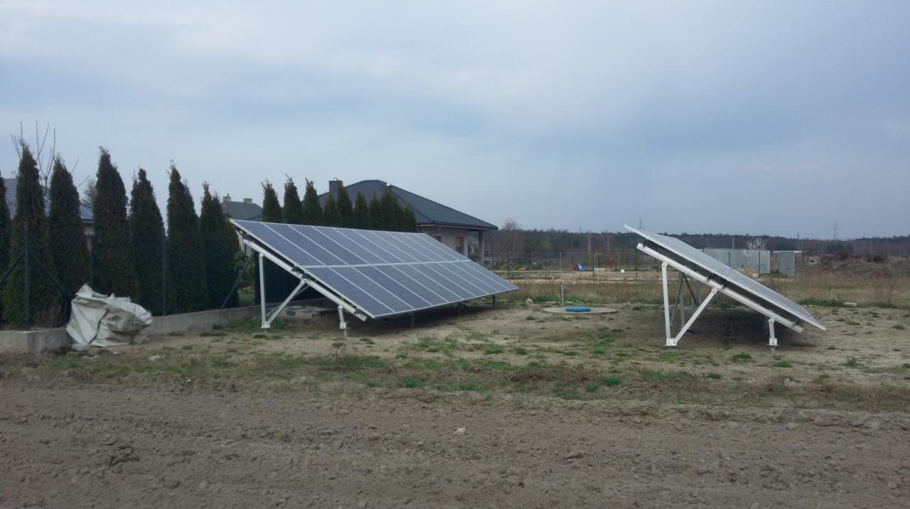 Dwa panele słoneczne zamontowane na ziemi przed domem jednorodzinnym, widoczne drzewa i pochmurne niebo.