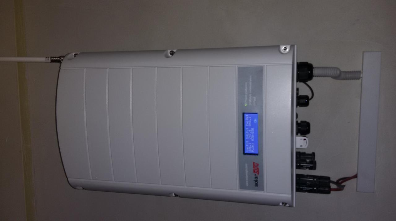 Szary inwerter SolarEdge zamontowany na ścianie z widocznymi kablami i wyświetlaczem pokazującym parametry pracy.