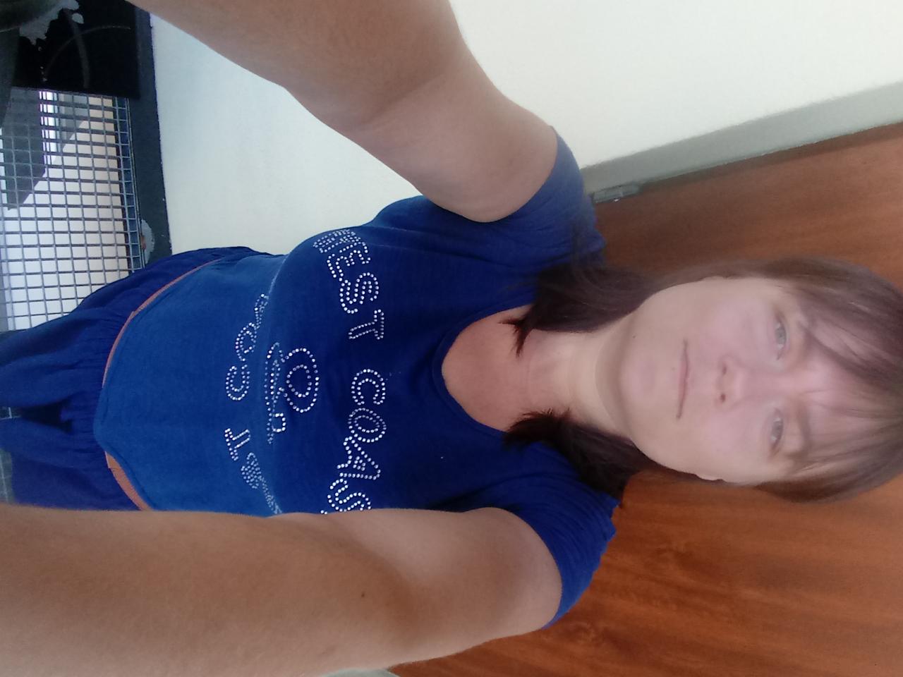 Kobieta w niebieskiej koszulce z napisem 'West Coast' pozuje w pomieszczeniu z kratką wentylacyjną i drewnianą ścianą.