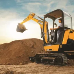 Żółto-czarna minikoparka JCB 30 Plus w trakcie pracy, operator w kabinie, na pierwszym planie pryzma ziemi, w tle budowa i zachmurzone niebo.