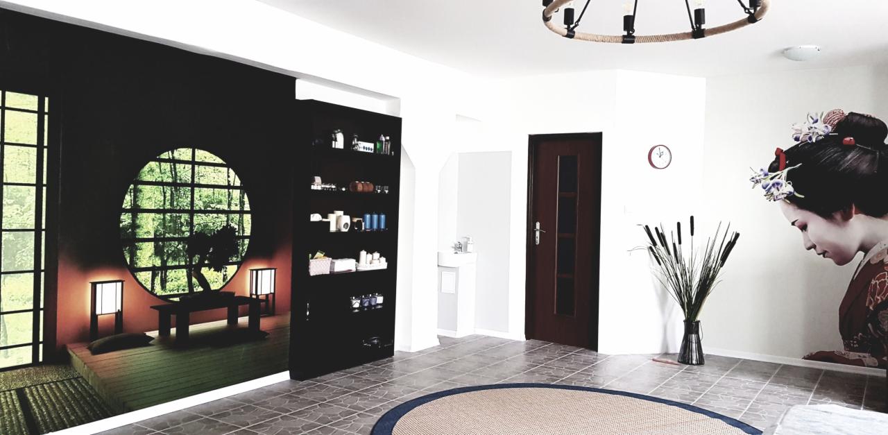 Wnętrze pokoju z elementami wystroju inspirowanymi stylem japońskim: fototapeta z motywem ogrodu zen i gejszy, okrągły dywan, lampa z jutowym sznurem, czarna półka z kosmetykami, trzciny w wazonie.