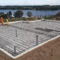 Zbrojenie płyty fundamentowej na piasku, widoczne rury kanalizacyjne, szalunek styropianowy, w tle jezioro i zalesione wzgórza.