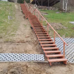 Długie, drewniane schody z poręczami prowadzące w górę po zboczu, obok dom na palach i dom z białym balkonem, w tle las.