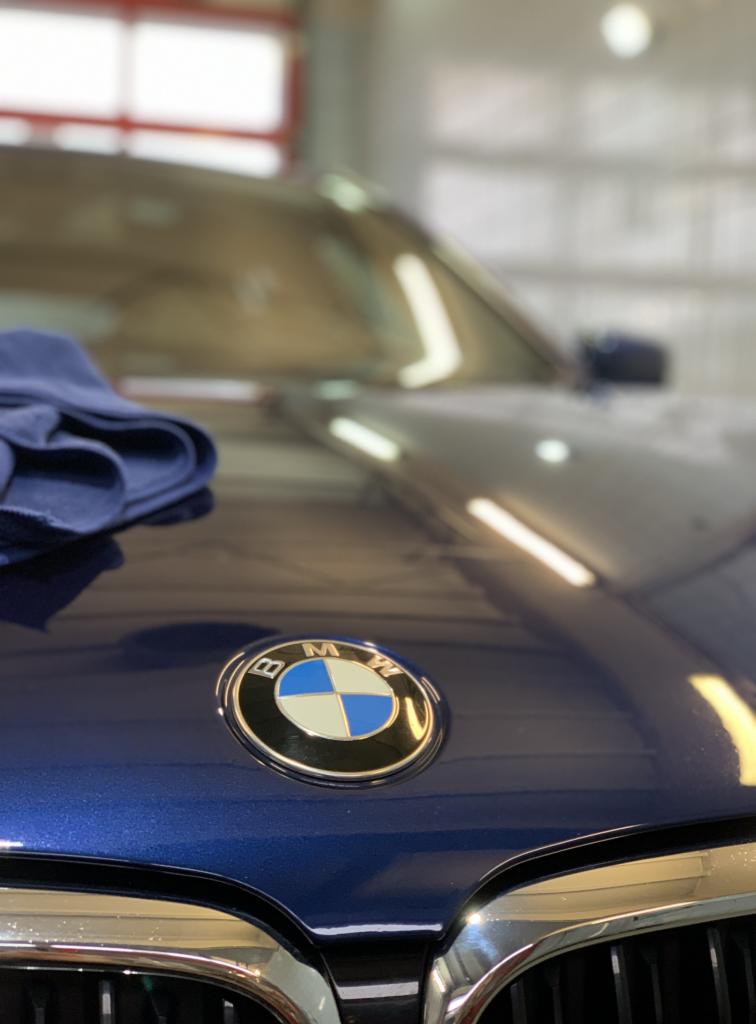Błyszcząca, granatowa maska samochodu BMW z widocznym logo i fragmentem grilla, na masce leży złożona niebieska ściereczka z mikrofibry, w tle rozmyte wnętrze myjni.