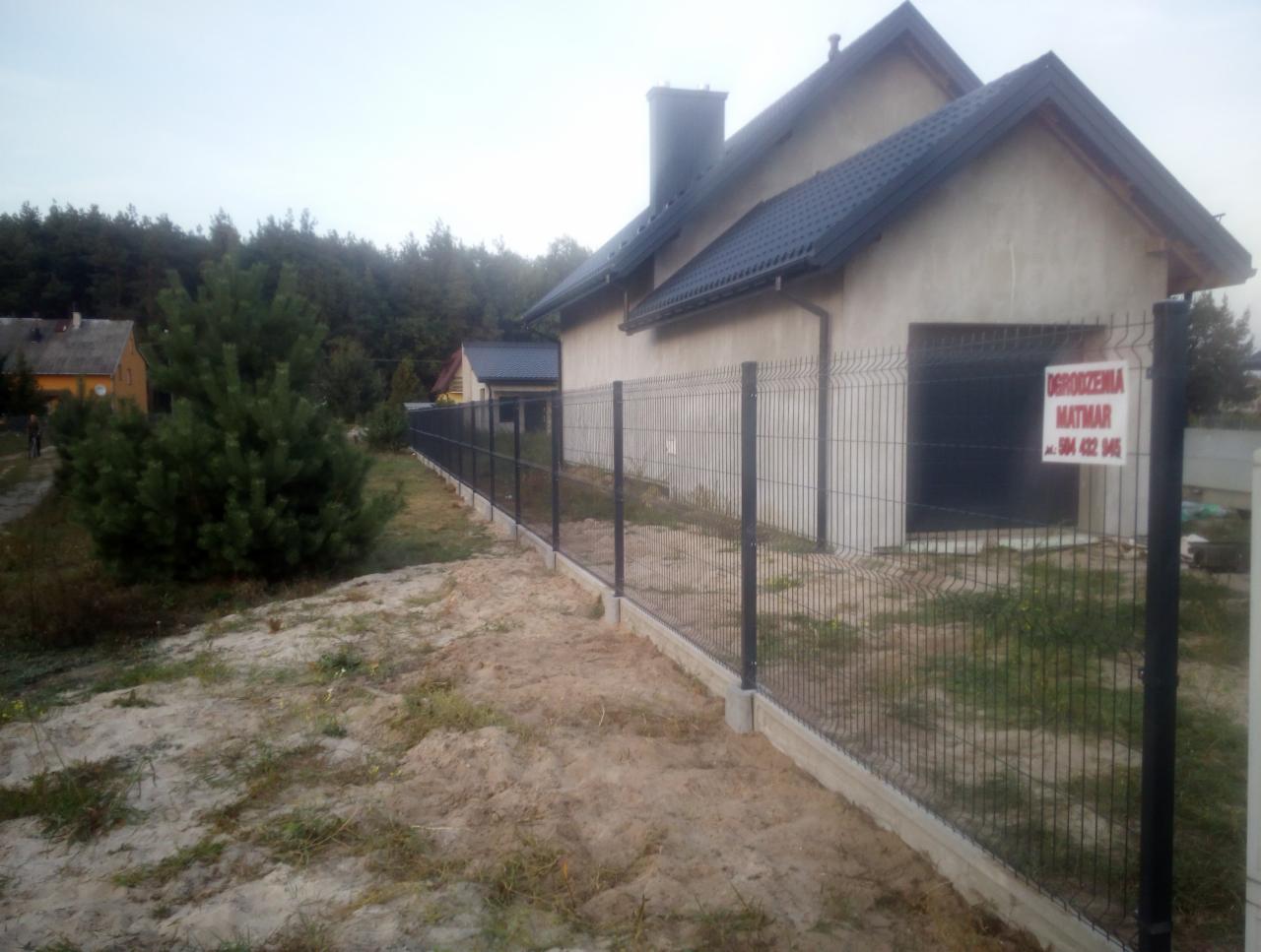 Nowoczesne, grafitowe ogrodzenie panelowe z podmurówką betonową otaczające posesję z domem jednorodzinnym i garażem; tabliczka reklamowa firmy na słupku.