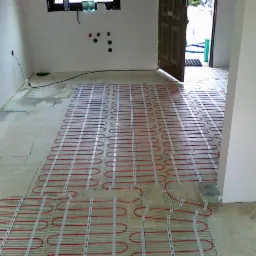 Rozłożona mata grzewcza z czerwonymi przewodami na podłodze w surowym stanie, widoczne otwory w ścianie pod instalację elektryczną i otwarte drzwi do innego pomieszczenia.