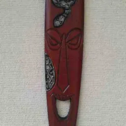 Podłużna, brązowa maska afrykańska z rzeźbioną twarzą, wężem u góry i metalowymi elementami dekoracyjnymi na policzku i dole.