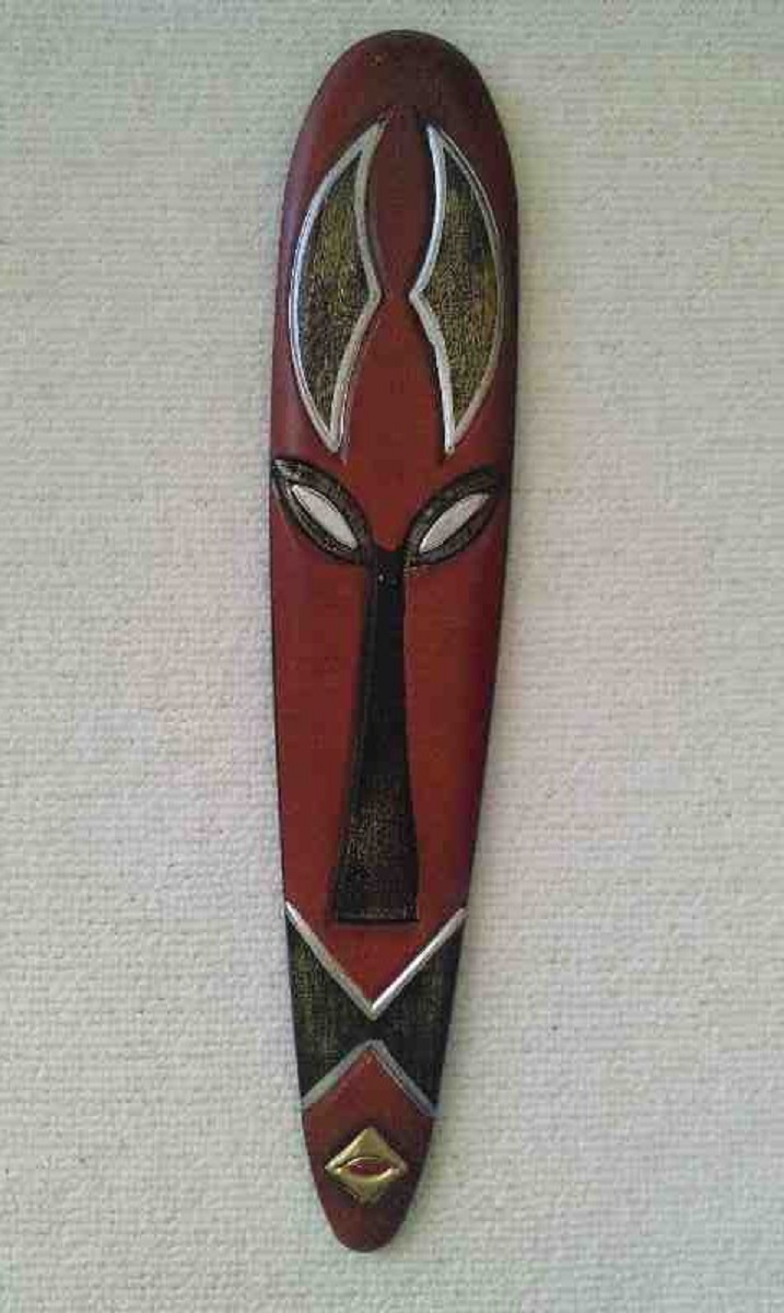 Podłużna, ręcznie malowana, dekoracyjna maska afrykańska w odcieniach czerwieni, czerni i złota, z geometrycznymi wzorami i srebrnymi akcentami, wisząca na teksturowanej, jasnej ścianie.