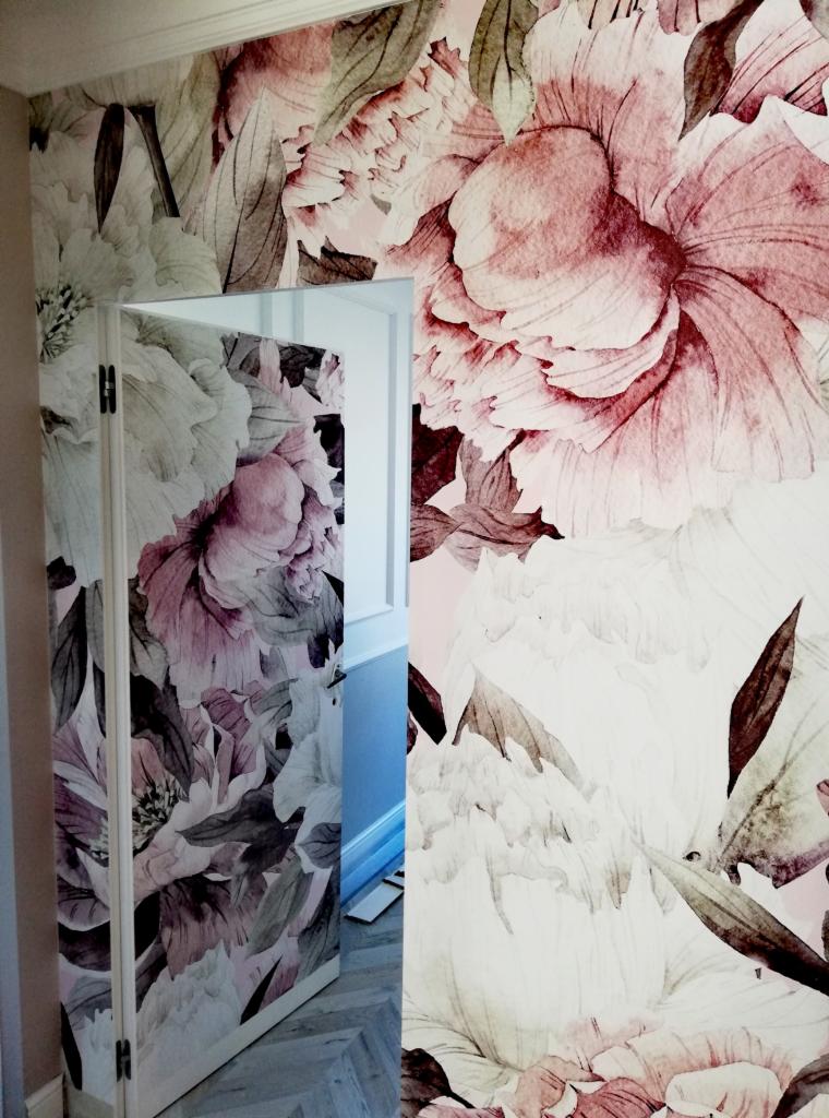 Wzorzysta tapeta w kwiaty zdobiąca ścianę i drzwi, z delikatnym motywem roślinnym w odcieniach różu i szarości, tworząca spójny i stylowy wystrój wnętrza.