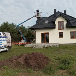 Pracownik na podnośniku koszowym czyści elewację domu jednorodzinnego za pomocą myjki ciśnieniowej; w tle zaparkowany van z logo firmy oferującej usługi mycia ciśnieniowego.