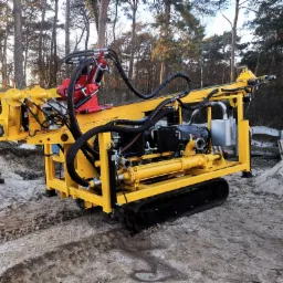 Żółta, gąsienicowa wiertnica na placu budowy z widocznymi przewodami hydraulicznymi i stertą piasku w tle.