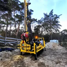 Żółta wiertnica na gąsienicach, z wysokim masztem wiertniczym, stoi na piaszczystym terenie z widocznymi śladami gąsienic; w tle las i przyczepa kempingowa.