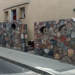 Kamienny mur ogrodzeniowy z dekoracyjnymi, metalowymi rozetami w okrągłych otworach, oddzielający posesję od chodnika i ulicy.