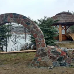 Kamienny łuk wejściowy z widokiem na jezioro i drewnianą altanę z trzcinowym dachem, fragment kamiennego murku, teren budowy w tle.