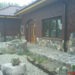 Murowany dom z elementami kamiennymi: podmurówka, ścieżka, obrzeża rabat. Widoczne okno, drzwi wejściowe, dachówka i roślinność. Detal: ceramiczna figurka kota.