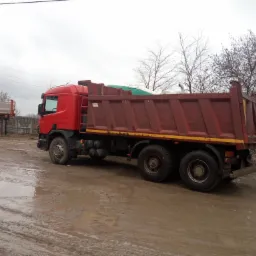 Czerwona wywrotka na błotnistym placu budowy, z widocznymi śladami opon i częściowo zakrytą zieloną plandeką w skrzyni ładunkowej.