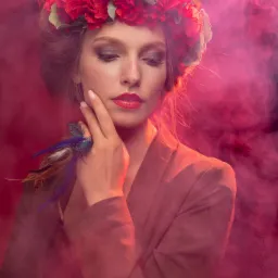 Portret kobiety w czerwonym wianku i marynarce, z pierścionkiem z piórami, w otoczeniu różowego dymu. Spojrzenie skierowane w dół, delikatny makijaż, artystyczna fotografia.