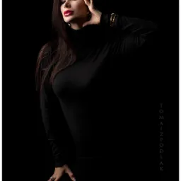 Portret kobiety w czarnym golfie na ciemnym tle, z mocnym makijażem ust i biżuterią, oświetlonej studyjnym światłem.