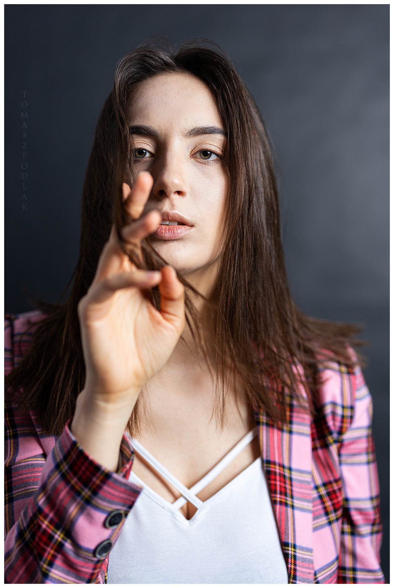 Portret kobiety z brązowymi włosami, w różowej kraciastej marynarce, trzymającej kosmyk włosów przed twarzą na ciemnoszarym tle.