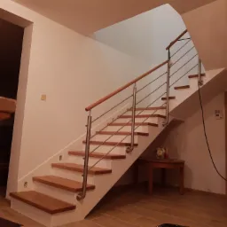 Nowoczesne schody z drewnianymi stopniami i metalową balustradą, widok od dołu, oświetlenie ciepłe, na podłodze płytki imitujące drewno.