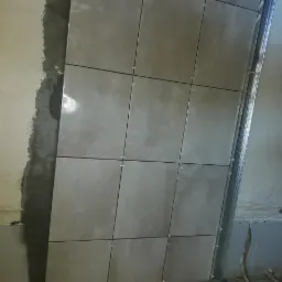 Świeżo ułożone, szare płytki na ścianie z widocznymi krzyżykami dystansowymi, obok fragment ściany przygotowanej do dalszego kładzenia płytek i metalowy profil.