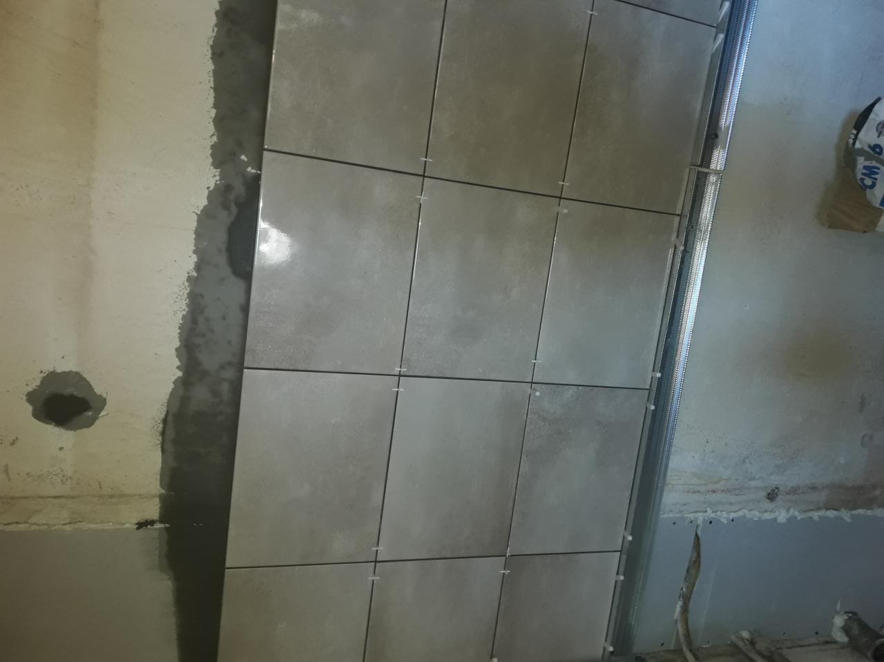 Świeżo ułożone, szare płytki na ścianie z widocznymi krzyżykami dystansowymi, obok fragment ściany przygotowanej do dalszego kładzenia płytek i metalowy profil.