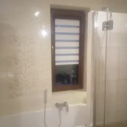 Wykończona łazienka z beżowymi płytkami ściennymi z delikatnym wzorem, białą wanną z brązową mozaiką na obudowie, oknem z roletą w drewnianej ramie i szklanym parawanem nawannowym.