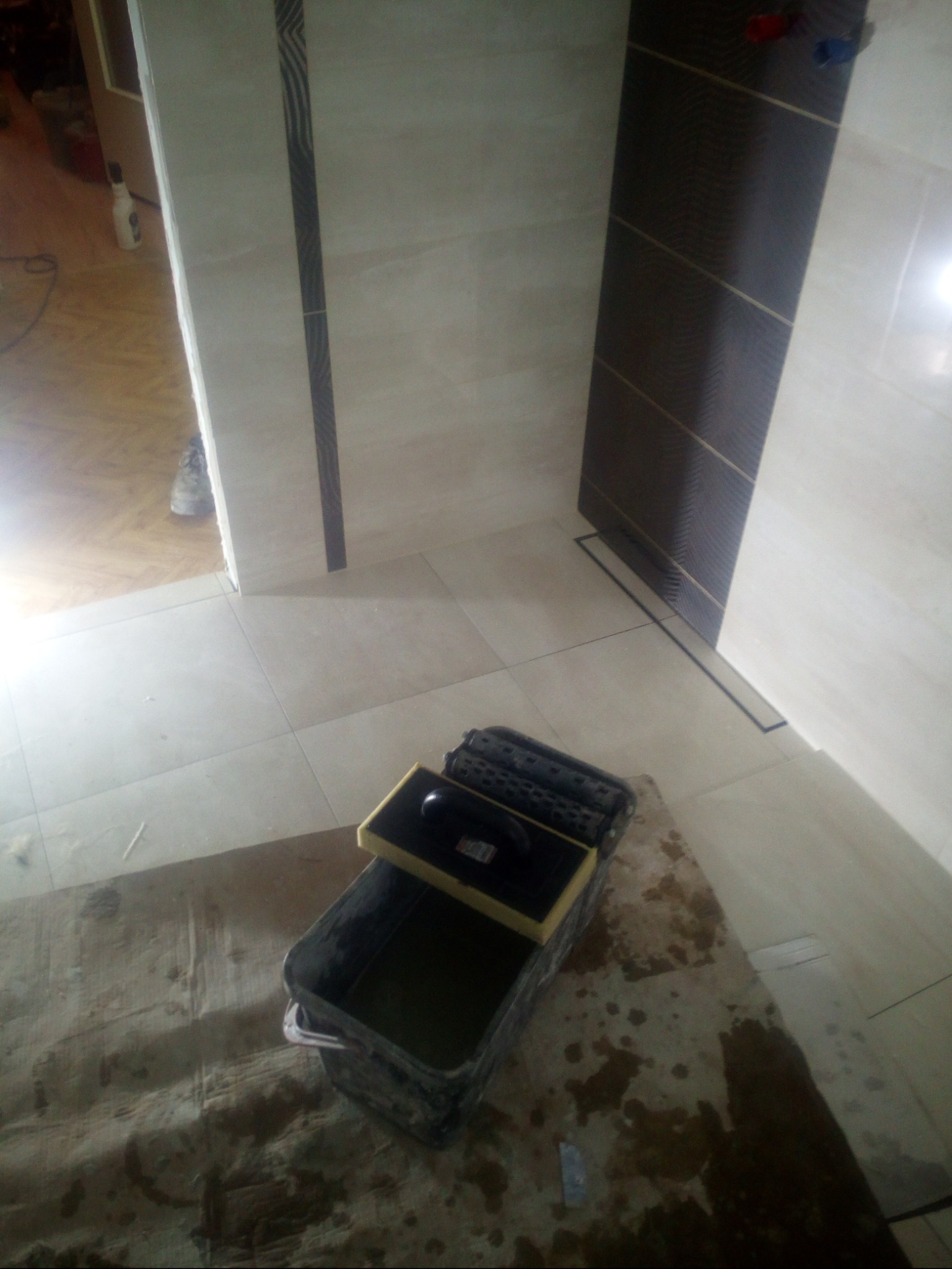 Wykończona łazienka z beżowymi płytkami i ciemnobrązowym akcentem, widoczny pojemnik z zaprawą i narzędzia do układania płytek na zabezpieczonej folią podłodze.