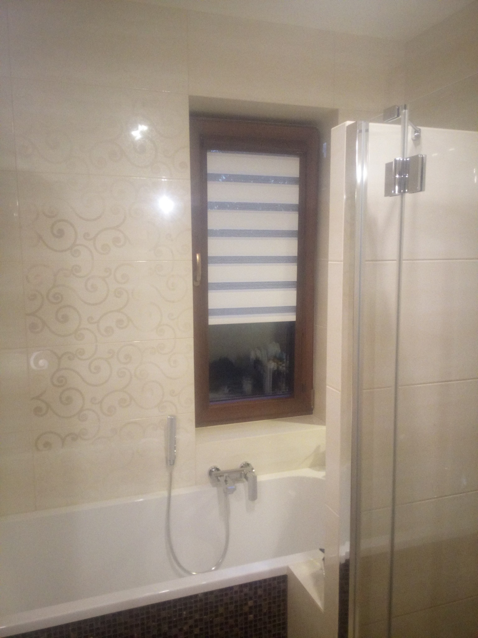 Wykończona łazienka z beżowymi płytkami ściennymi z delikatnym wzorem, białą wanną z brązową mozaiką na obudowie, oknem z roletą w drewnianej ramie i szklanym parawanem nawannowym.