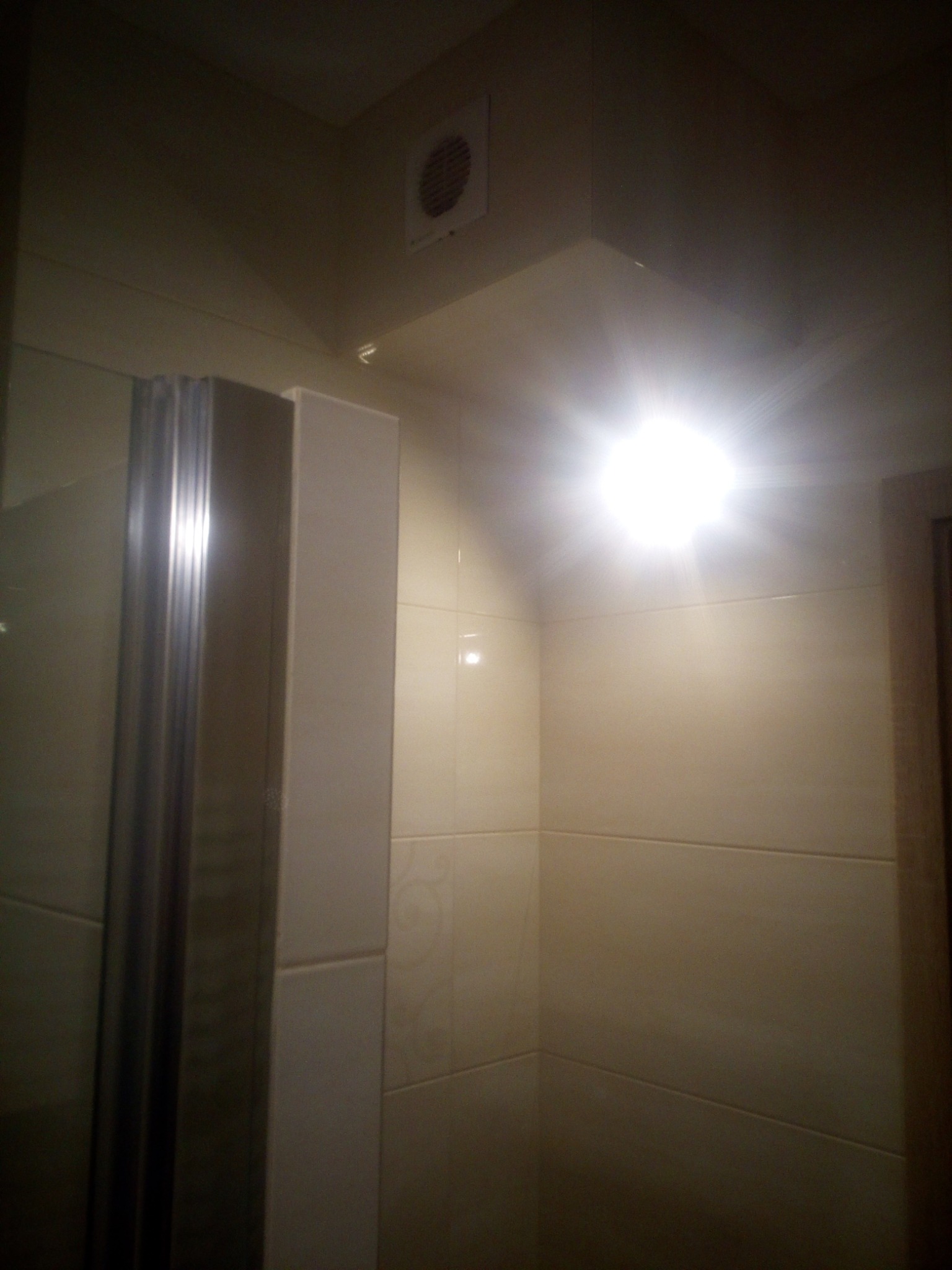 Wykończona łazienka z beżowymi płytkami, kabiną prysznicową z metalowymi profilami i widoczną kratką wentylacyjną pod sufitem, oświetlona mocnym światłem.