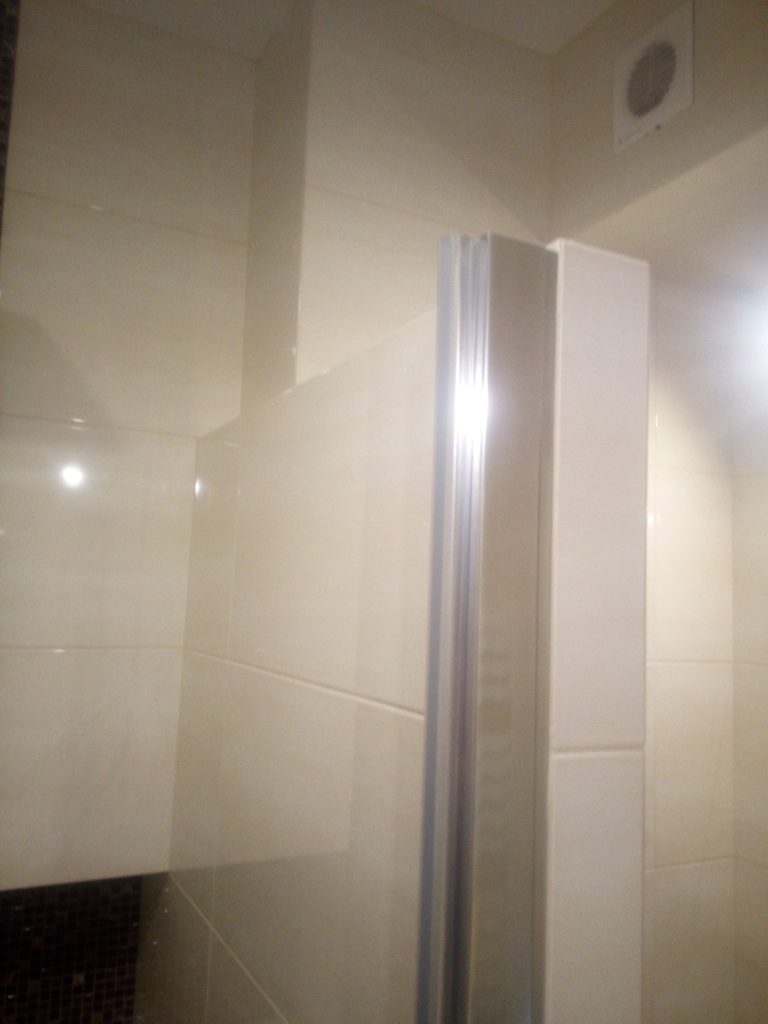 Jasne, kremowe płytki w kabinie prysznicowej z aluminiową ramą, widoczny fragment ciemnej mozaiki pod prysznicem i kratka wentylacyjna na suficie.