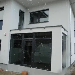 Nowoczesny dom z białymi ścianami i czarnymi oknami PCV oraz przeszklonymi drzwiami przesuwnymi, widoczne krople deszczu, częściowo widoczny samochód dostawczy.