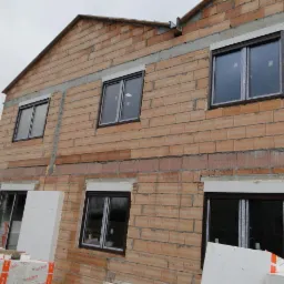 Surowy stan budynku w trakcie budowy z zamontowanymi oknami z brązowymi ramami i częściowo opuszczonymi białymi roletami zewnętrznymi; widoczne bloki styropianu.