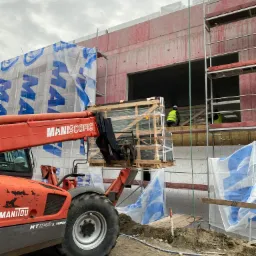 Montaż okien na budowie: podnośnik Manitou transportuje skrzynię z oknami na rusztowanie, gdzie pracownicy w kamizelkach odblaskowych przygotowują się do instalacji.