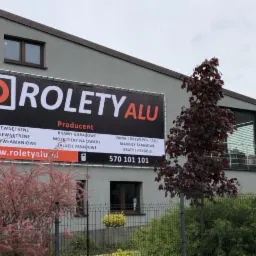 Budynek firmy produkującej rolety, bramy garażowe i okna, z dużym banerem reklamowym na elewacji, prezentującym logo i ofertę, widoczne okna i rolety w budynku, otoczenie z zielenią.